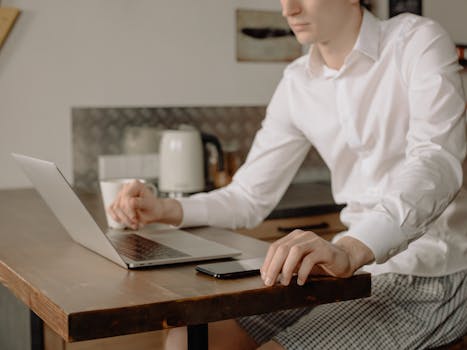 A man in casual attire works on a laptop in his kitchen, embracing remote work life.