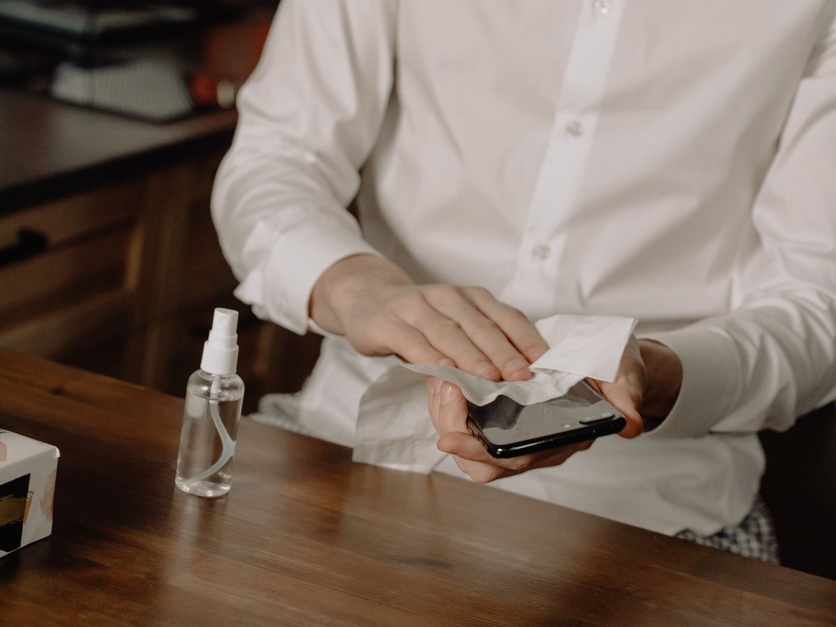 Person wiping a smartphone at a table with a spray bottle and tissues nearby