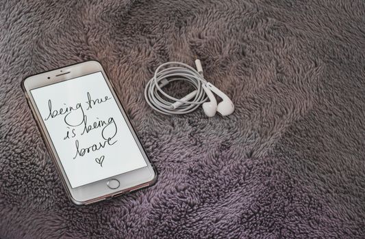 Smartphone displaying 'being true is being brave', with earphones, on a cozy fluffy blanket.