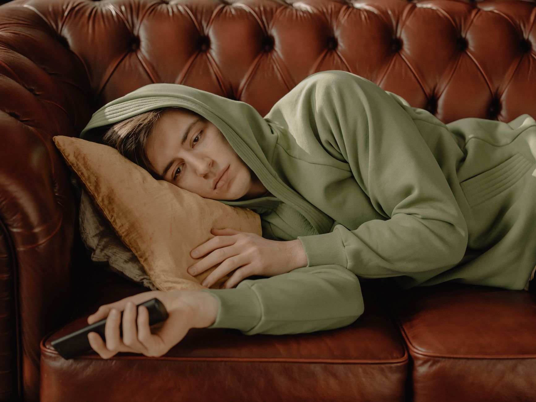 A young man in a green hoodie lying on a leather sofa at home with a remote control.