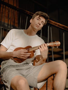 Young man playing ukulele at home, enjoying a relaxed moment in sunlight.