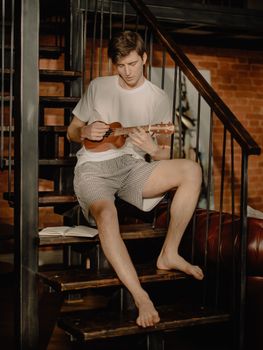 Casual young man playing ukulele on stairs inside a cozy home.