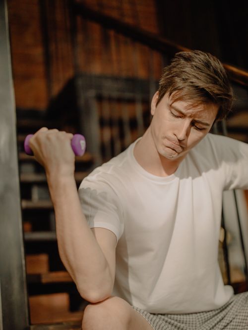 Free A young man in a white t-shirt performs an exercise with a dumbbell indoors. Stock Photo