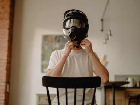 Man in white t-shirt fastening a motorcycle helmet indoors with cozy home interior.