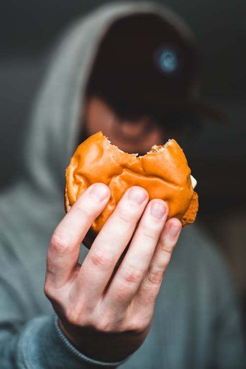 Selective focus of anonymous male in casual clothing and hat with bitten burger