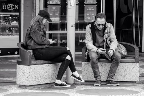 Free People Sitting on Benches in front of Shops in a City and Scrolling Through Their Smartphones Stock Photo