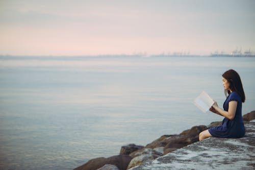 Free A Woman Sitting Beside the Ocean while Reading Book Stock Photo