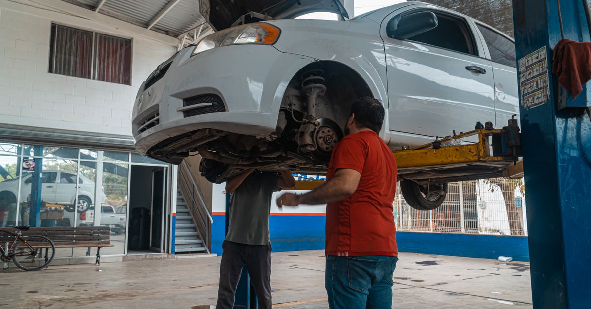 Two mechanics inspecting a white car on a lift inside a garage workshop.