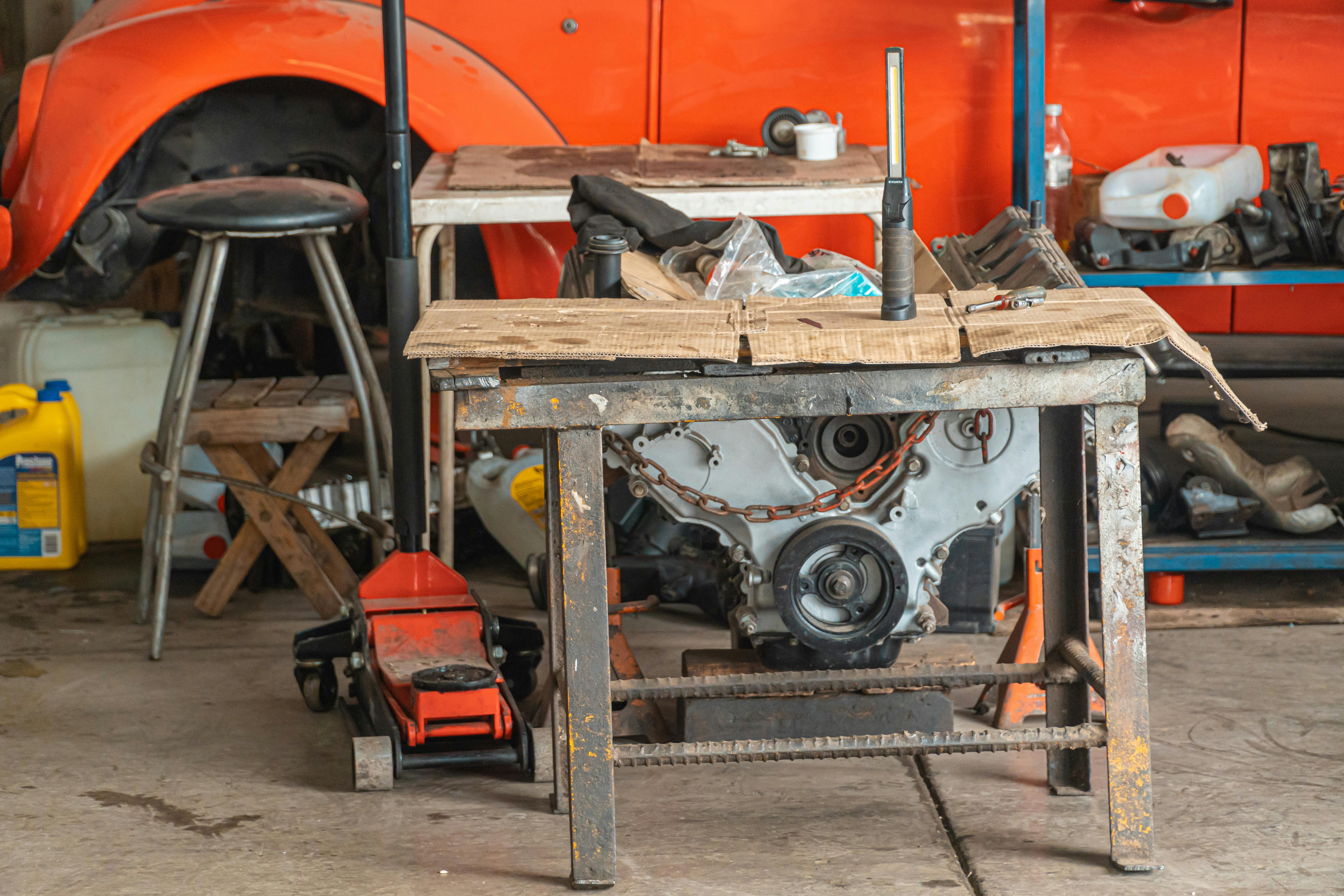 Old car engine on a workshop table with tools and a red car in the background.