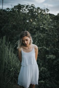 一位身着白色连衣裙的女子，静静地站在户外，四周环绕着郁郁葱葱的绿植