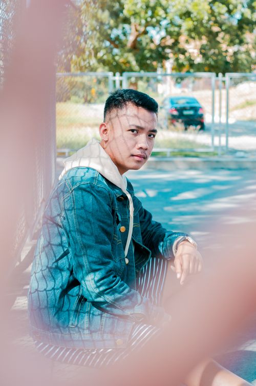 Free Side view of casual Asian male in denim jacket looking at camera sitting on metal fence on street Stock Photo