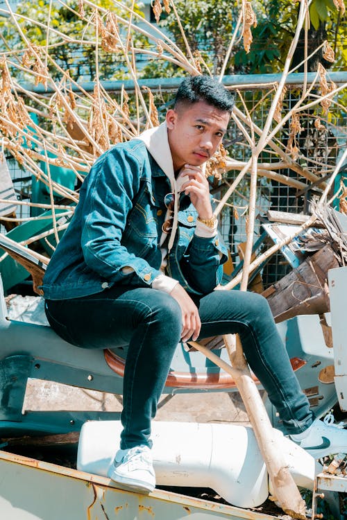 Free Confident Asian man in sneakers and denim clothes touching chin while looking at camera sitting in local poor dock Stock Photo