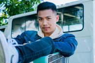 Modern Asian man in denim shirt and sneakers sitting in metal push cart looking at camera in yard