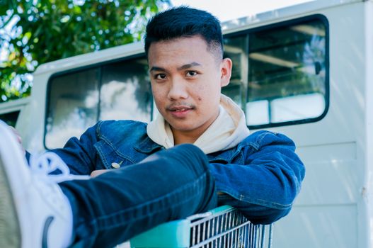 Modern Asian man in denim shirt and sneakers sitting in metal push cart looking at camera in yard