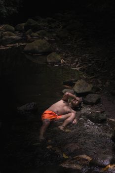 一个只穿着橙色短裤的男人在溪流边的岩石旁放松