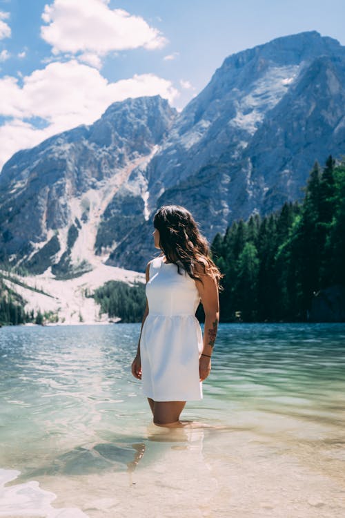 Woman in White Dress Standing on Water Near Snow Covered Mountain