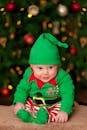 穿着绿色精灵服装的婴儿微笑着坐在装饰好的圣诞树前，捕捉节日的欢乐