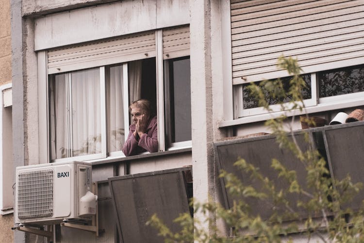 An Elderly Woman Looking Out Of An Apartment Window