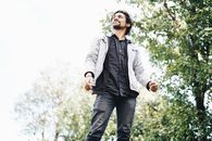一个穿着夹克、留着梦幻大胡子的男人站在街边的绿树下，望着远处