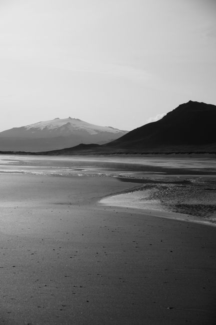 Black and white smooth sandy beach with calm sea waves and dark hills on background