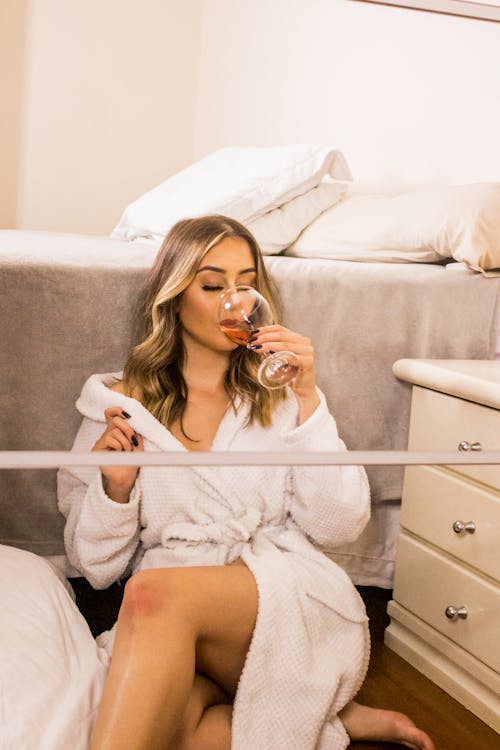 Sensual woman in white bathrobe sitting at bed in front of mirror and sipping red wine in leisure