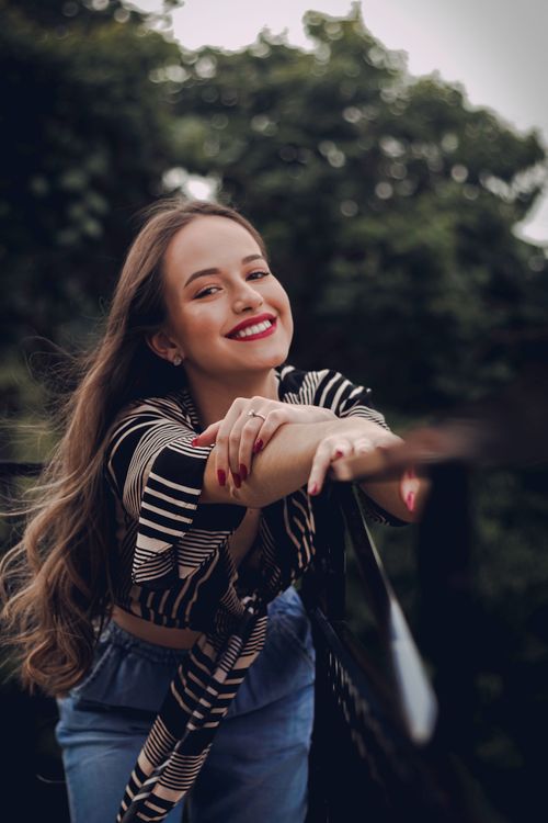 Free Charming young female with long waving hair and red lips smiling at camera leaning on fence in summer park Stock Photo