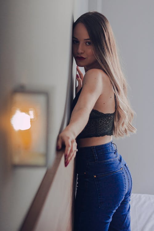 Side view of casual brunette in denim and lace top standing close to wall looking alluringly at camera