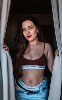 Alluring young woman in stylish sportive underwear and unbuttoned denim looking at camera standing in curtains
