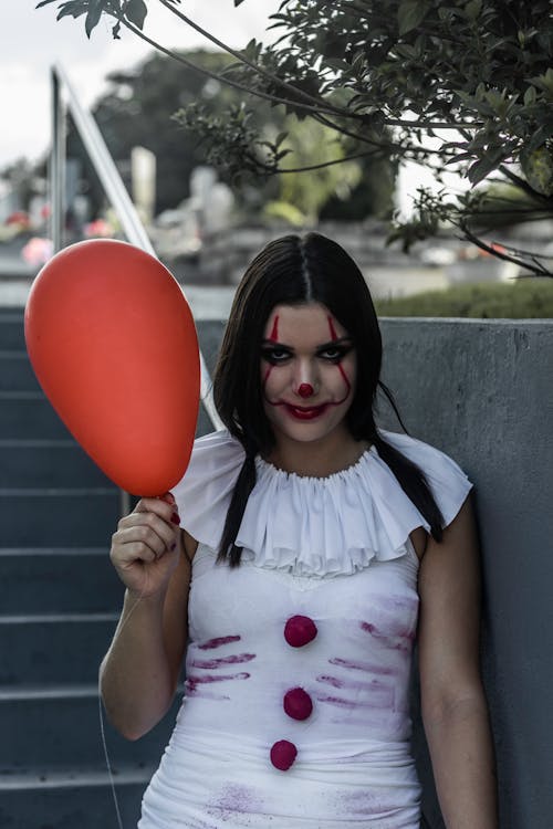 Young woman wearing spooky Halloween costume of clown with red balloon standing on street