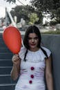 一个穿着怪异万圣节服装的年轻女子，扮成小丑拿着红气球站在街上