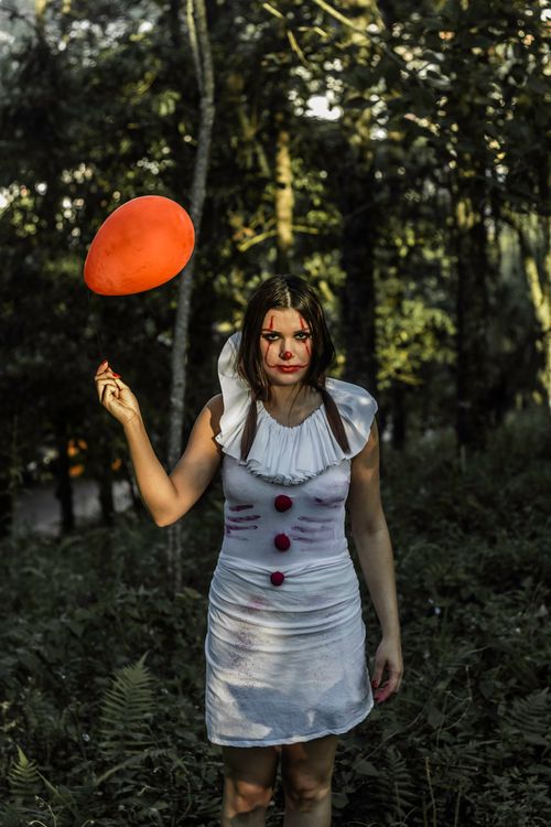 Free From above of fearful young woman in white costume and with clown makeup holding red balloon while standing in green park looking at camera Stock Photo