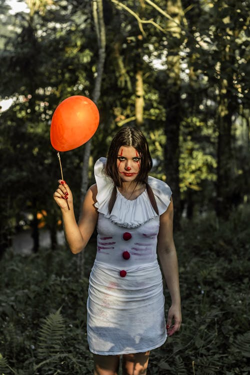 High angle of monstrous woman in white dress and with clown makeup holding red balloon standing in forest