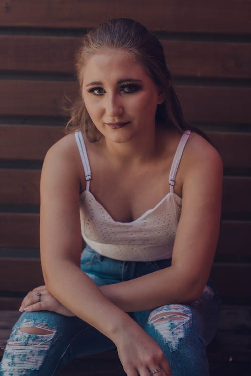 Free Young female in white crop top with bare shoulders sitting on wooden floor and looking at camera Stock Photo