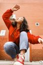 Low angle of trendy ethnic lady in jeans and bright red jacket squatting on cement floor with raised hand and closed eyes near brick wall in daylight