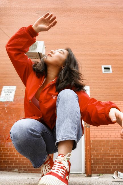 Low angle of trendy ethnic lady in jeans and bright red jacket squatting on cement floor with raised hand and closed eyes near brick wall in daylight