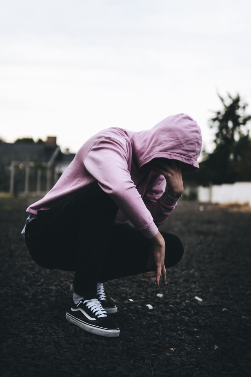 A person crouches outdoors in a pink hoodie, creating a moody silhouette. Urban street vibe.