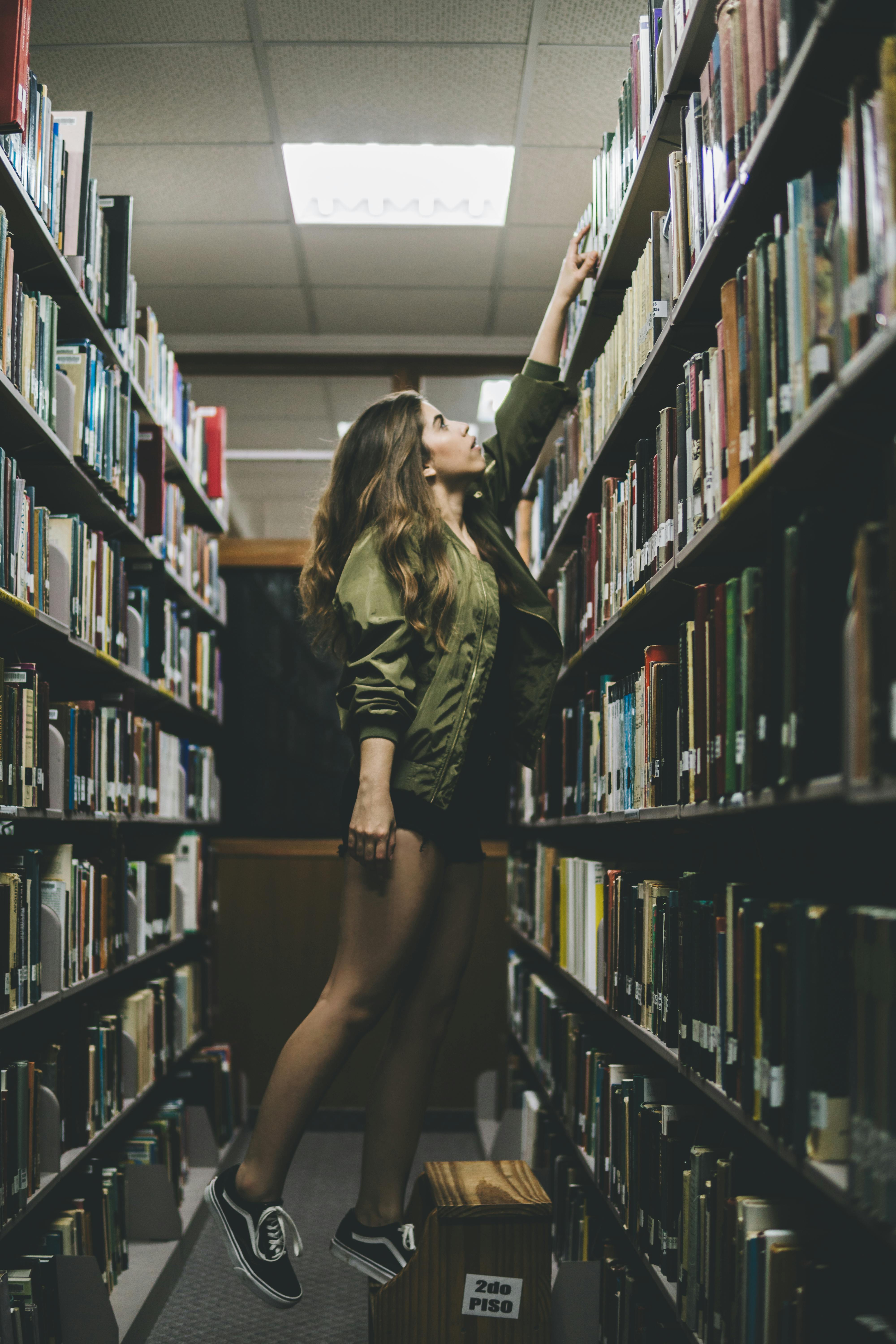 a woman reaching for a book on top of a bookshelf