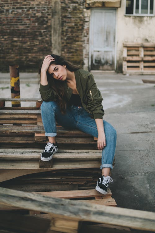 Woman in Green Long Sleeve Shirt and Blue Denim Jeans Sitting on Brown Wooden Stairs