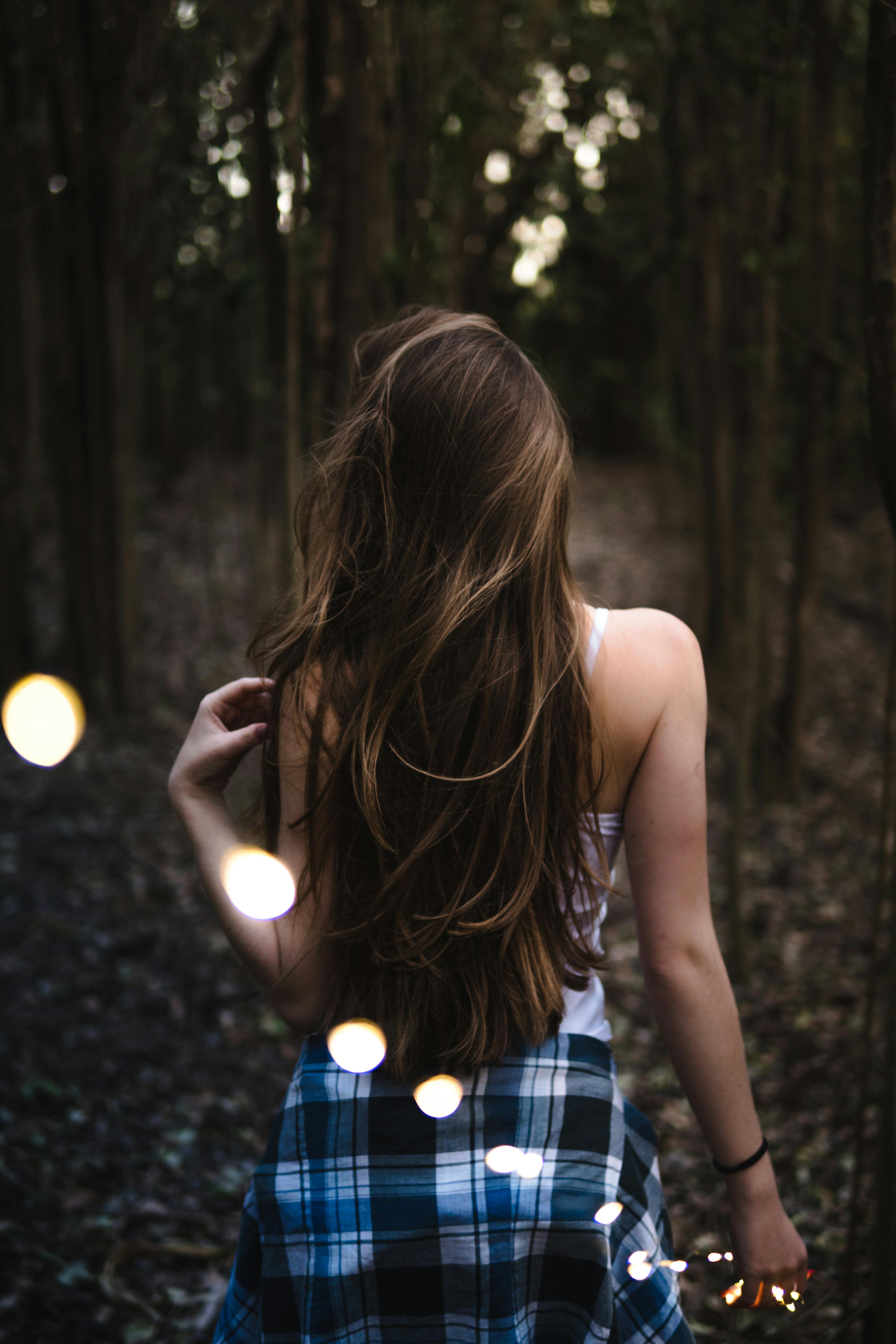 Back view of a woman walking in a forest holding string lights, creating a moody atmosphere.