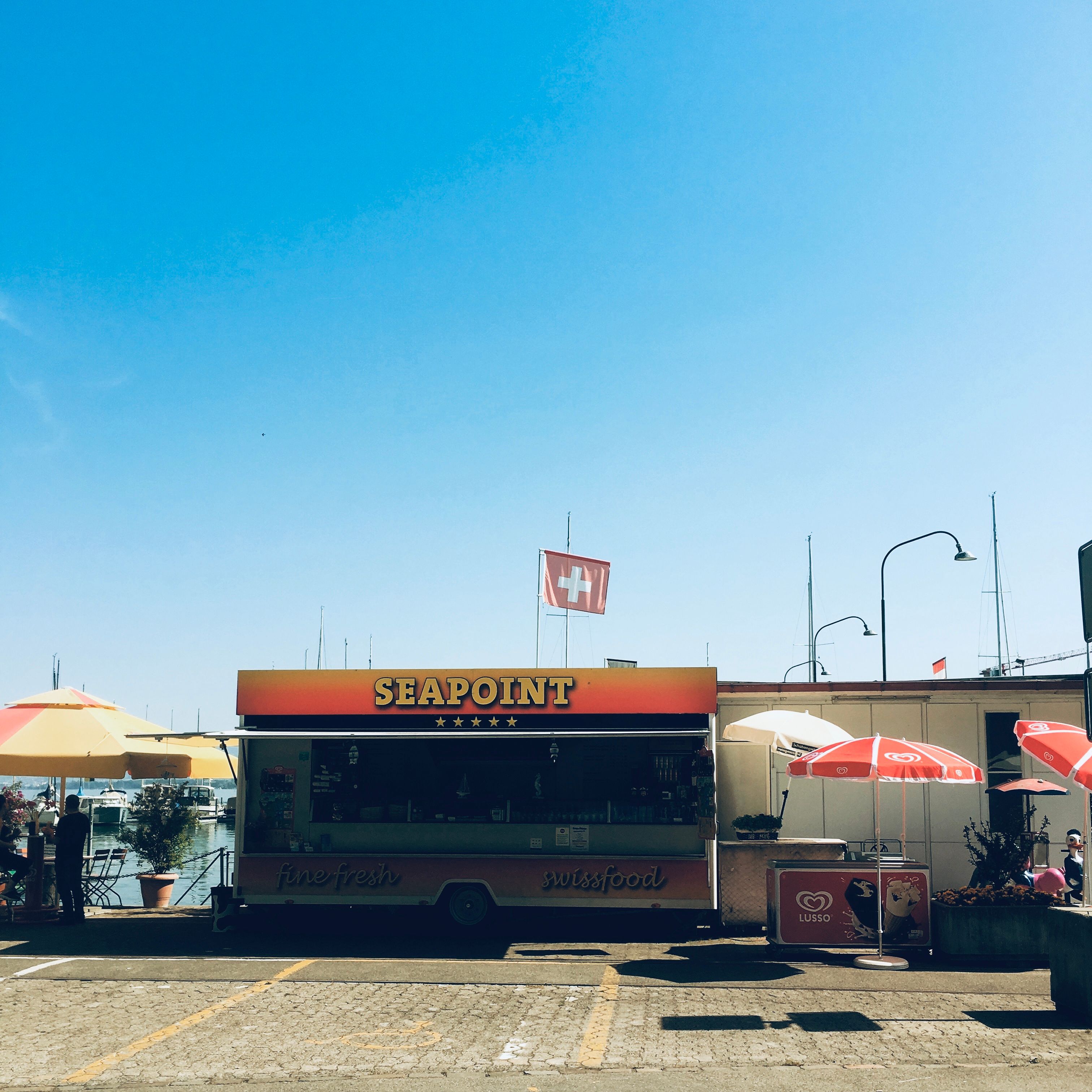 Street food van with umbrellas on paved street and flag Switzerland above with background of city harbor in sunny day