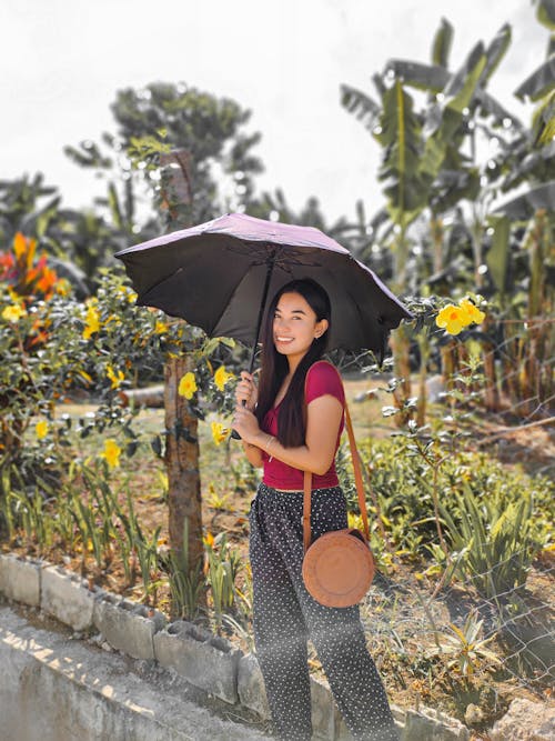 Positive Asian female tourist with umbrella covering from sunshine standing near fenced park with exotic plants and palms