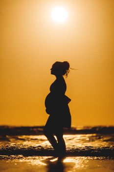 Silhouette of a pregnant woman on a beach during a vibrant sunset reflecting on the water.