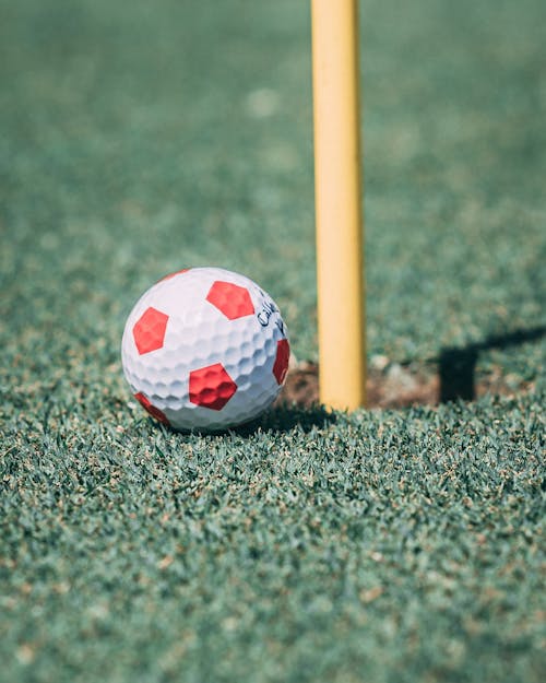 From above of small colorful golf ball with ribbed surface and pentagon ornament on meadow with artificial grass near vertical stick