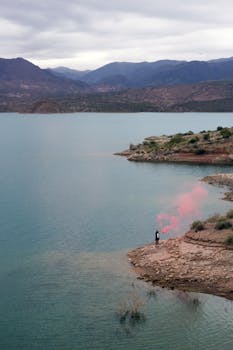Lone person by Argentina lake with pink smoke, mountainous landscape, dramatic ambiance.