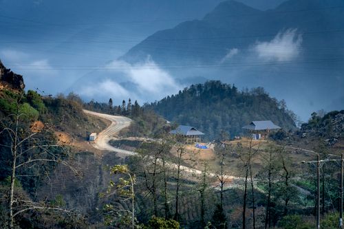 Free Narrow road going through green forest near mountains and small houses under blue sky Stock Photo