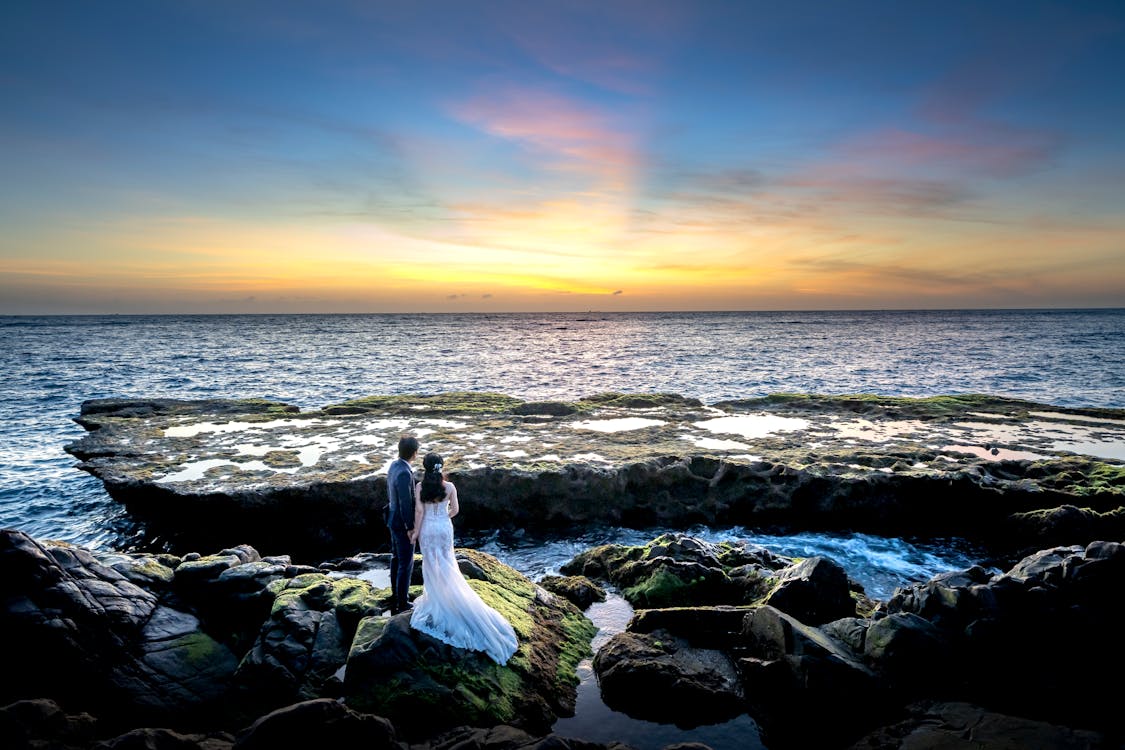 Photo of Couple Standing on Rocky Shore During Golden Hour