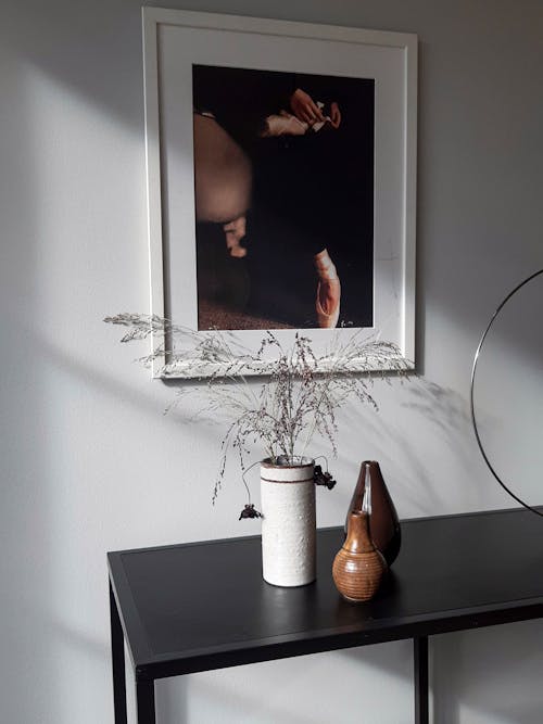 Black table with different vases placed near gray wall with picture in daylight