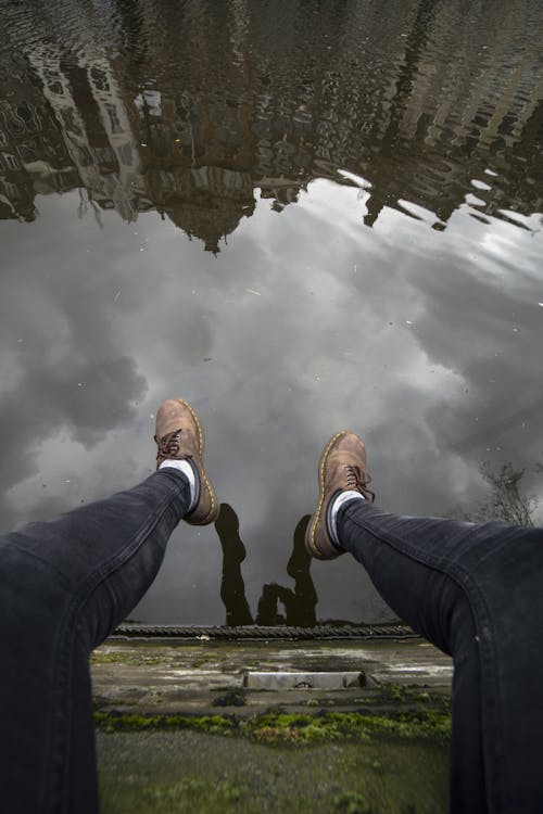 From above crop anonymous male in casual clothes resting on mossy stone canal front edge and hanging legs down over muddy water with reflection of sky and buildings