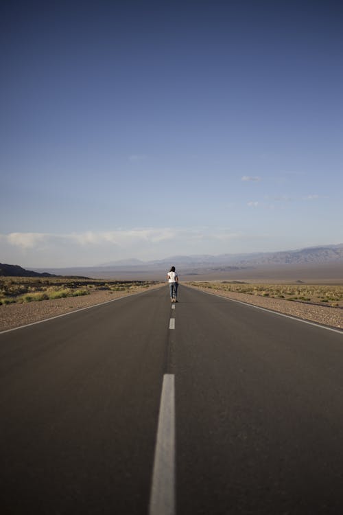 Back view full body faceless person in casual wear standing in center of empty remote asphalt road in countryside against rocky highlands on clear day