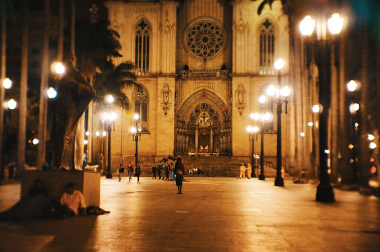 Facade Of Ancient Catholic Church Located On Square With Lanterns And Palm Trees At Night